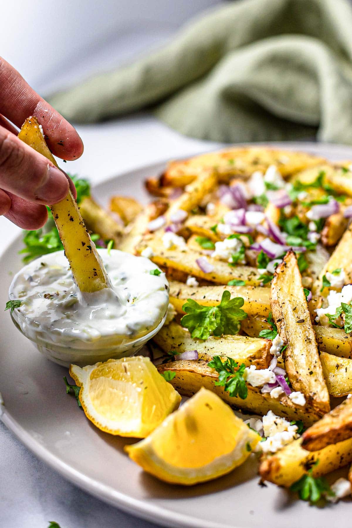 hand dipping greek fry into tzatziki with plate of fries beside.
