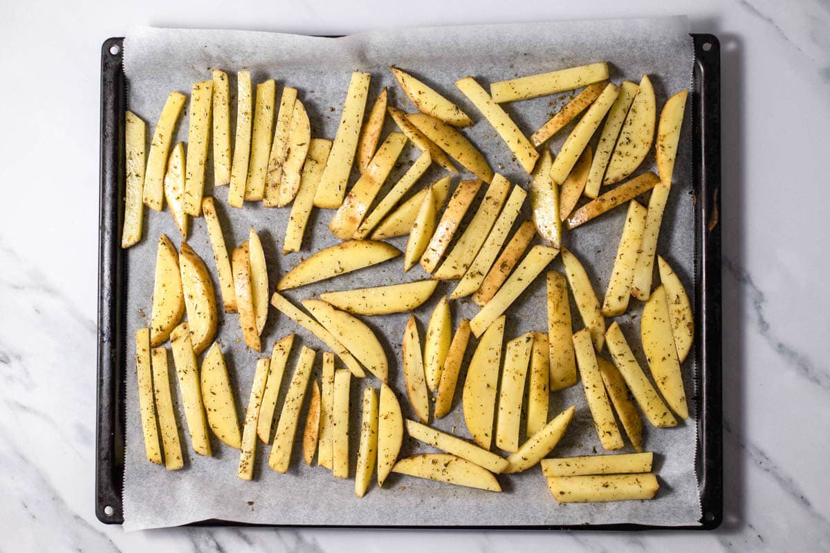baking sheet covered in parchment paper with raw greek fries laying on top.