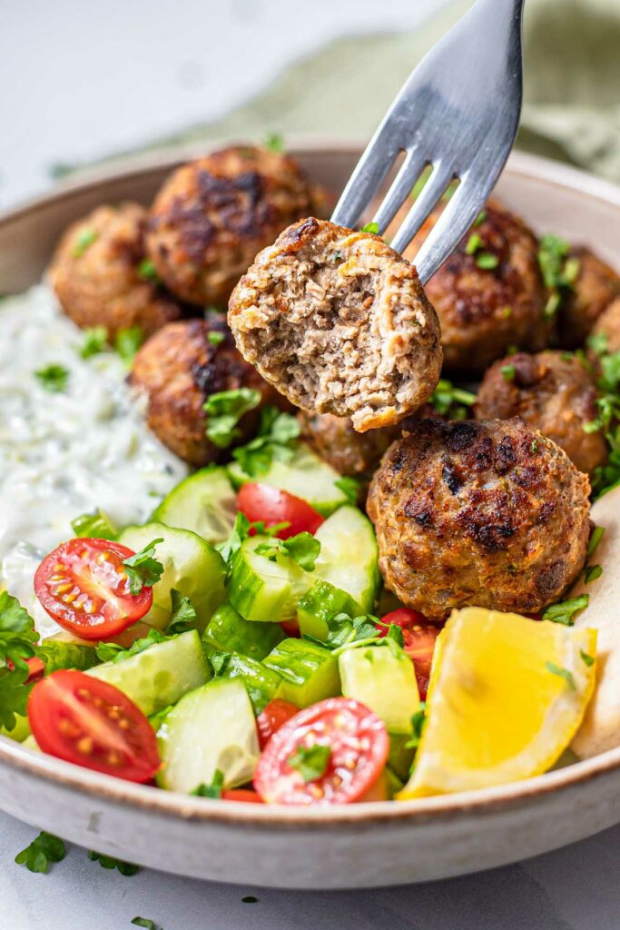 greek meatball on fork with bite out of it above plate of greek food.