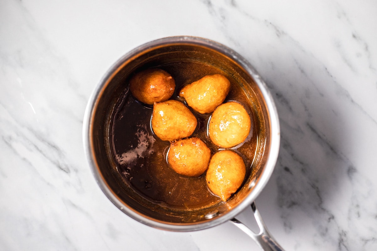 fried greek donuts floating in silver pot fill of honey mixture sitting on marble counter.