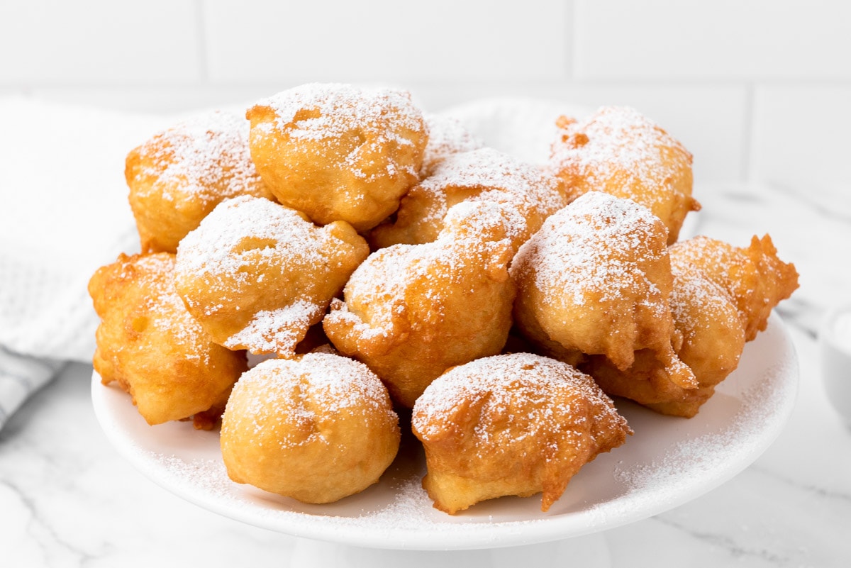 fried donut balls covered in powdered sugar sitting on white platter.