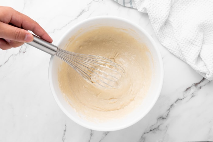 hand holding silver whisk mixing donut dough in white mixing bowl.