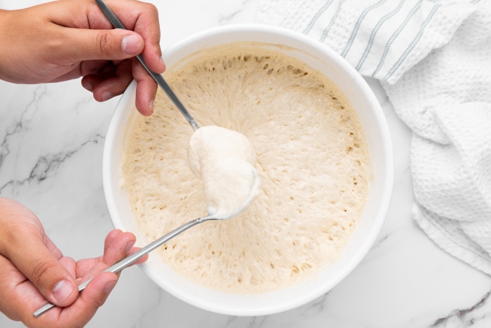 hands with two spoons dipping into doughnut batter in large bowl.