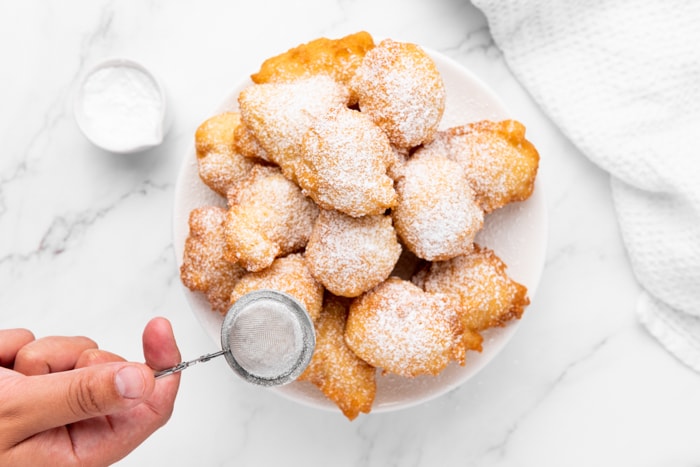 cooked dutch donuts on plate being covered in powdered sugar.