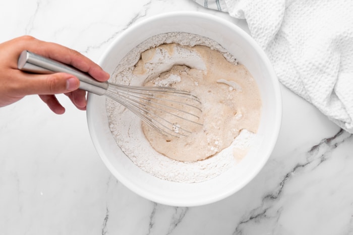 hand holding whisk mixing ingredients in white mixing bowl on marble counter.