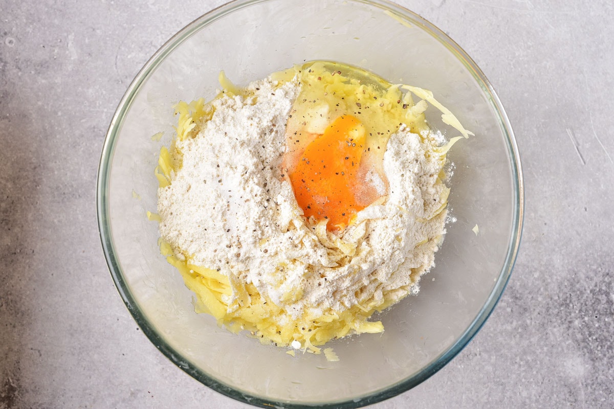 raw egg sitting in well of flour and shredded potato in clear glass bowl on counter top.