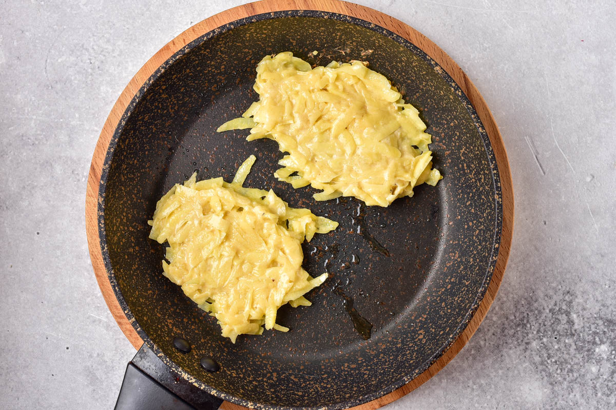 raw potato pancakes frying in oil in black frying pan with grey counter around.