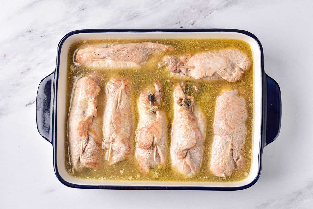 seven cooked pork rolls in large baking pan with yellow broth in it sitting on counter top.