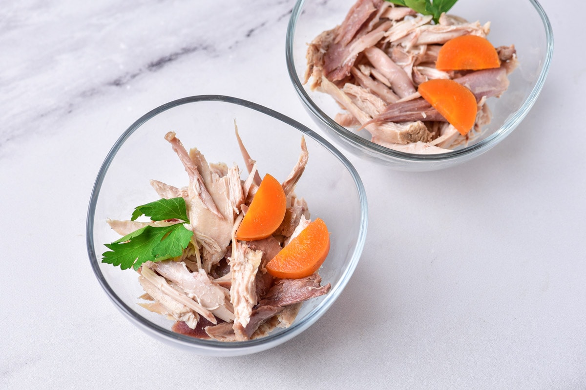 two clear bowls of cooked chicken and carrots with parsley sitting on white counter.