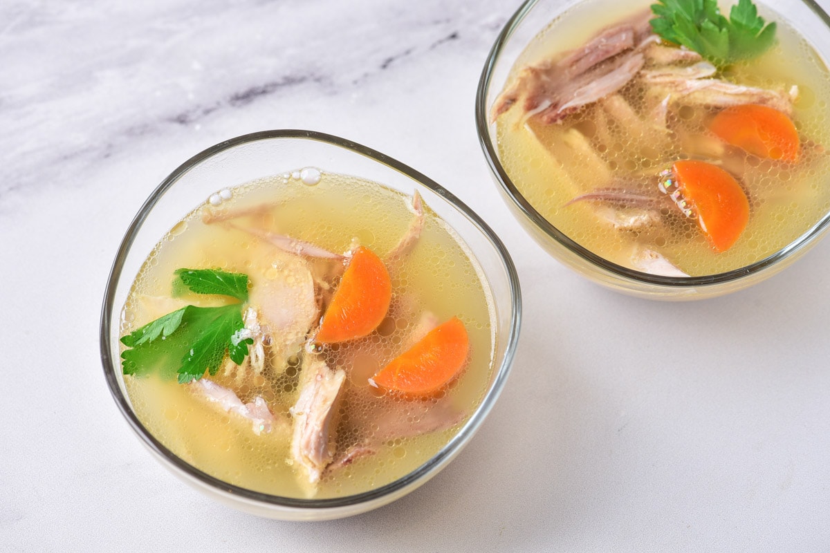 two clear glass bowls of cooked chicken and carrots filled with broth sitting on counter top.