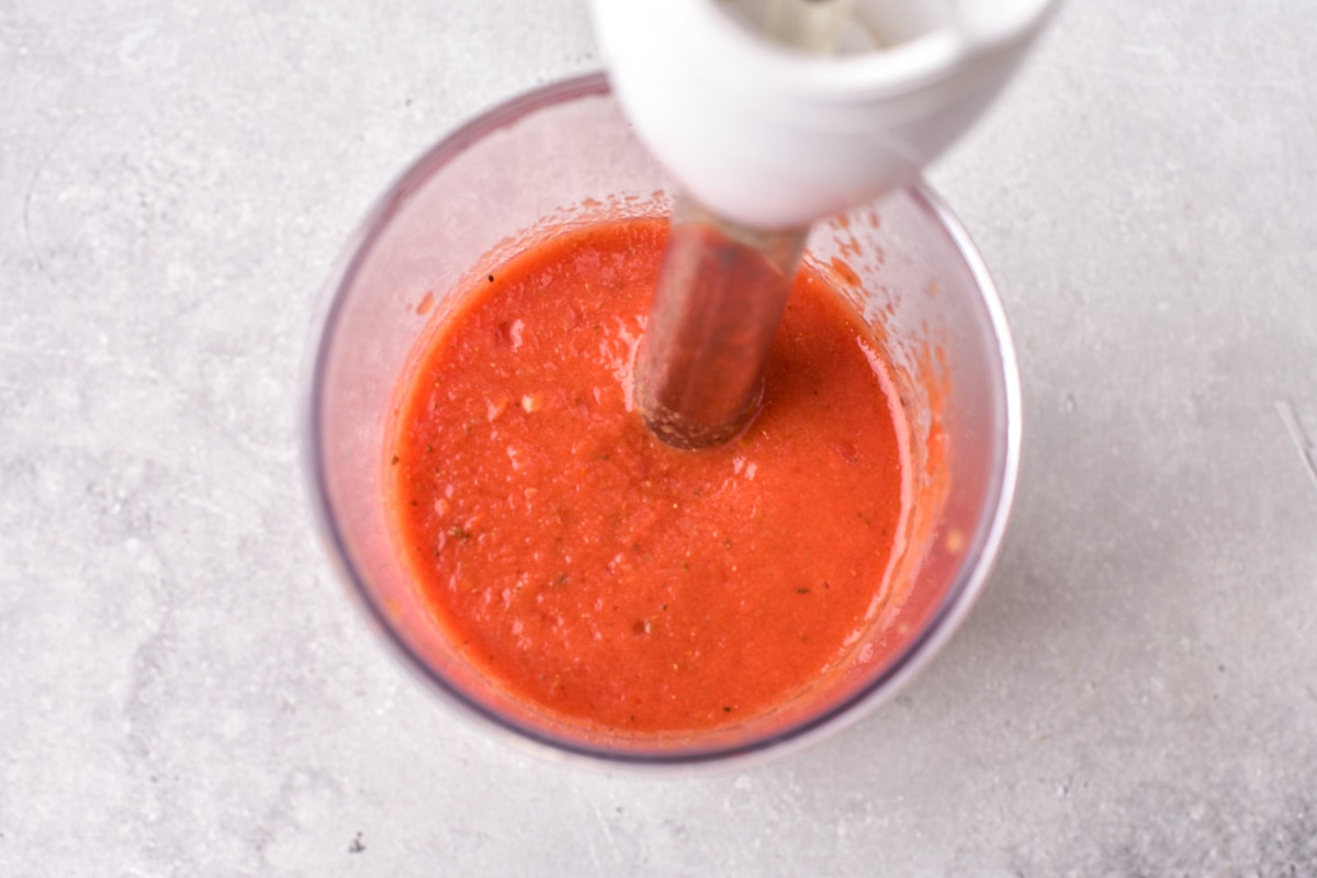 immersion blender mixing red tomatoes in glass dish on counter.