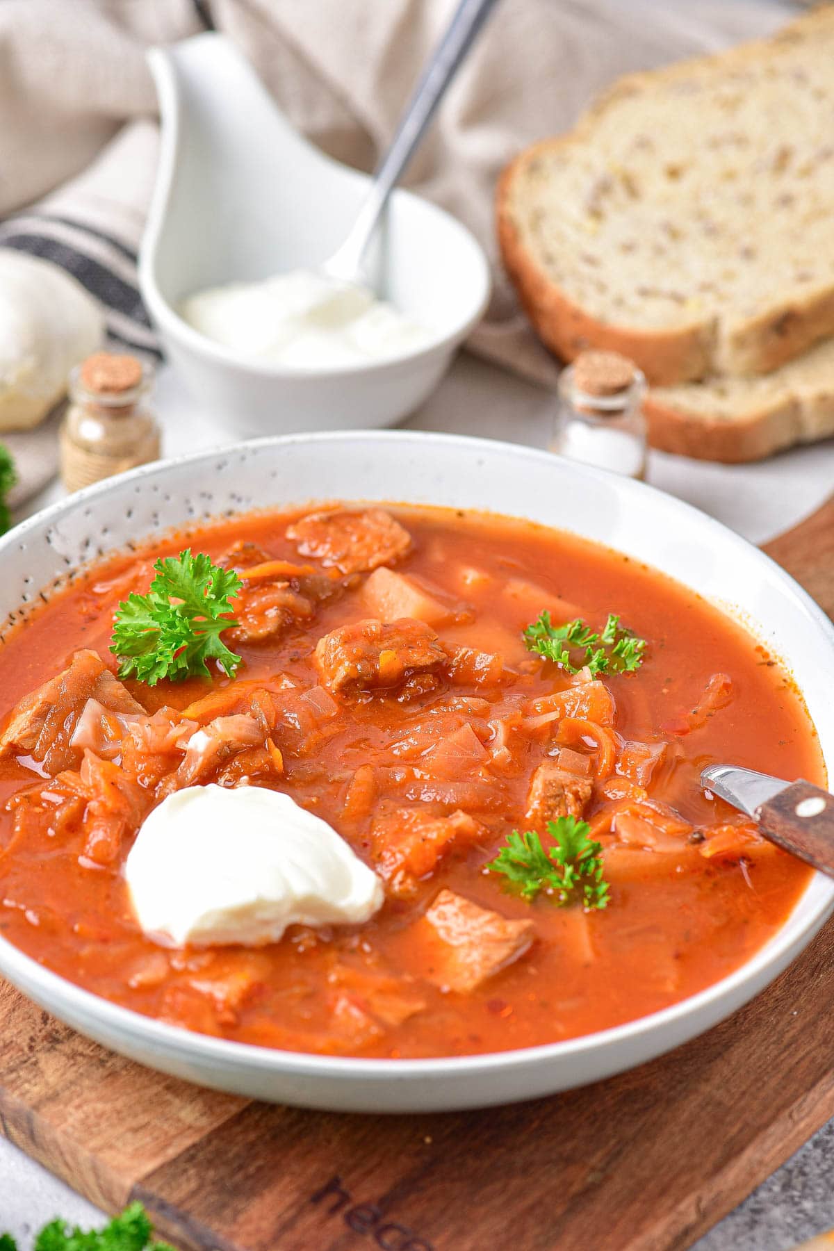 bowl of red borscht soup on wooden board with sour cream on top and slices of bread behind.