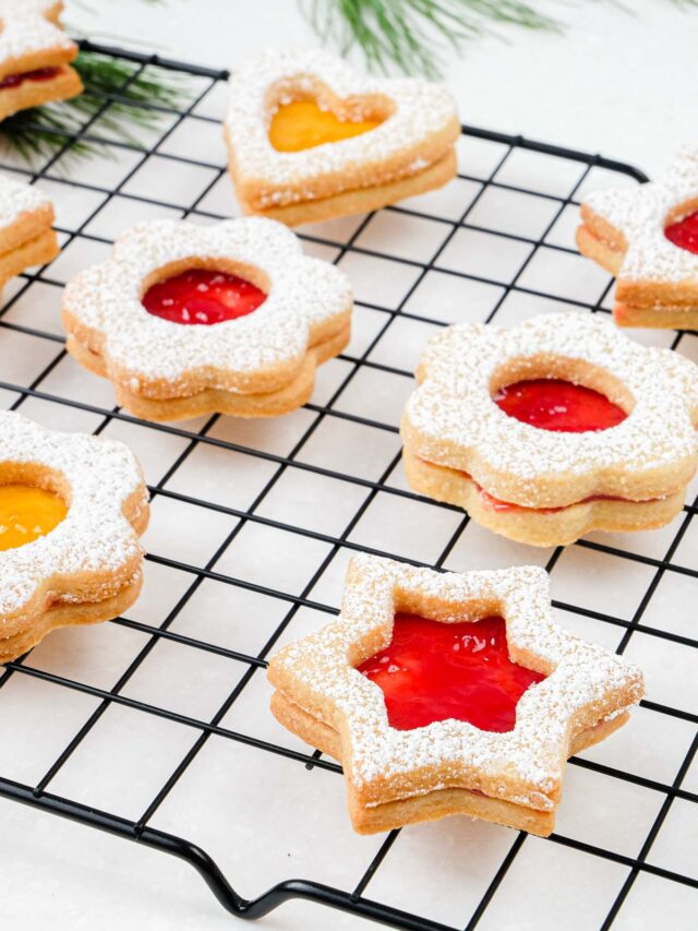 jam filled linzer cookies shaped like stars and flowers cooling on black wire cooking rack on white counter.
