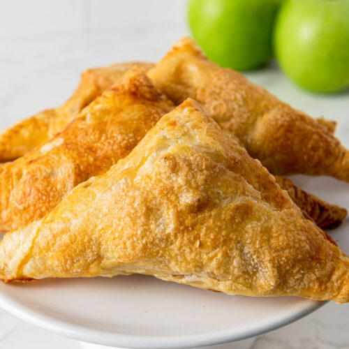flakey apple turnovers on white plate in white counter with green apples behind.