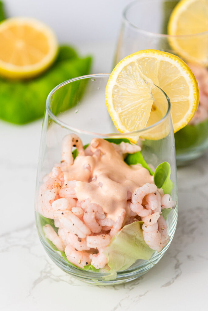 serving glass with cooked shrimp and creamy sauce on a bed of lettuce with slice of lemon on the rim.