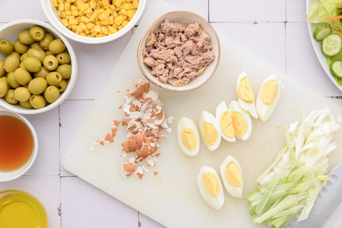 sliced hard boiled egg and pieces of canned tuna on white cutting board with other salad ingredients beside.