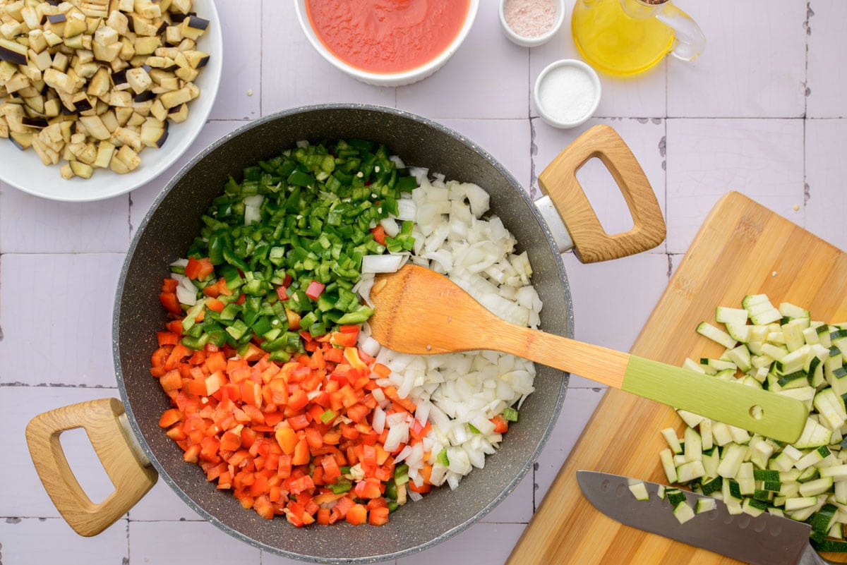 diced vegetables in black cast iron pan with wooden spoon hanging out beside cutting board and other vegetables.