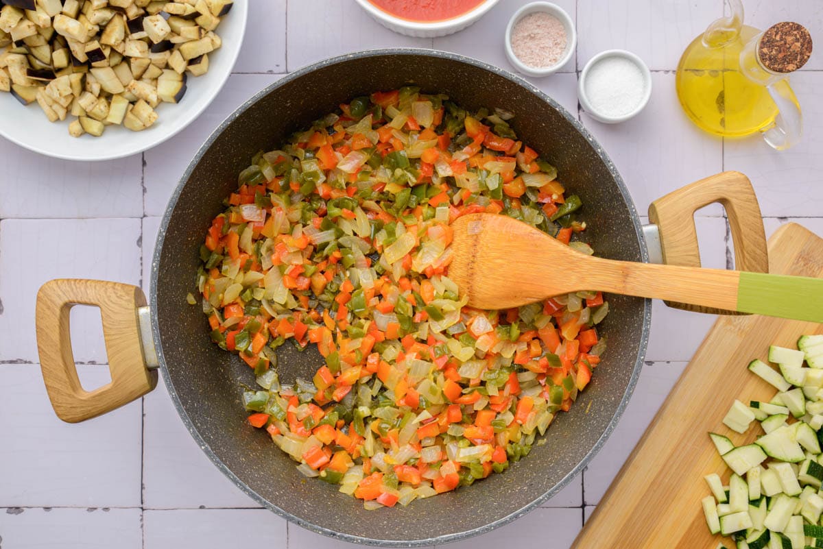 diced vegetables frying in black pan with wooden spoon and other ingredients around.