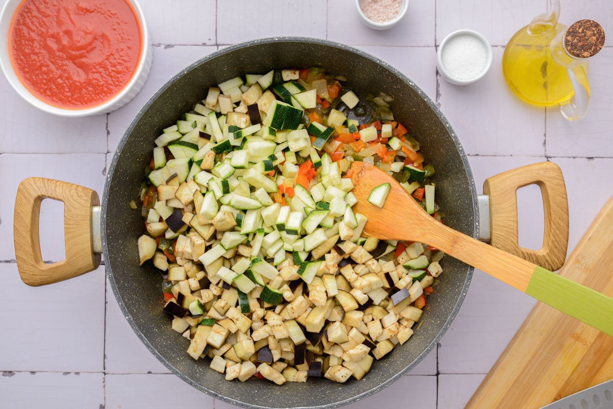 raw diced vegetables added to black pan of cooked vegetables with wooden spoon.