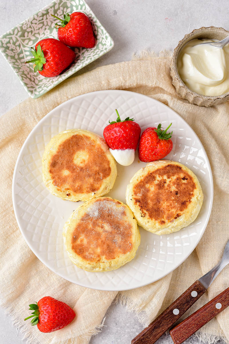 three golden brown cheese pancakes on white plate with whole strawberries dipped in cream beside.