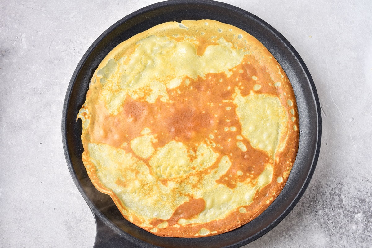 fried crepe with golden brown top in black frying pan with grey counter around.