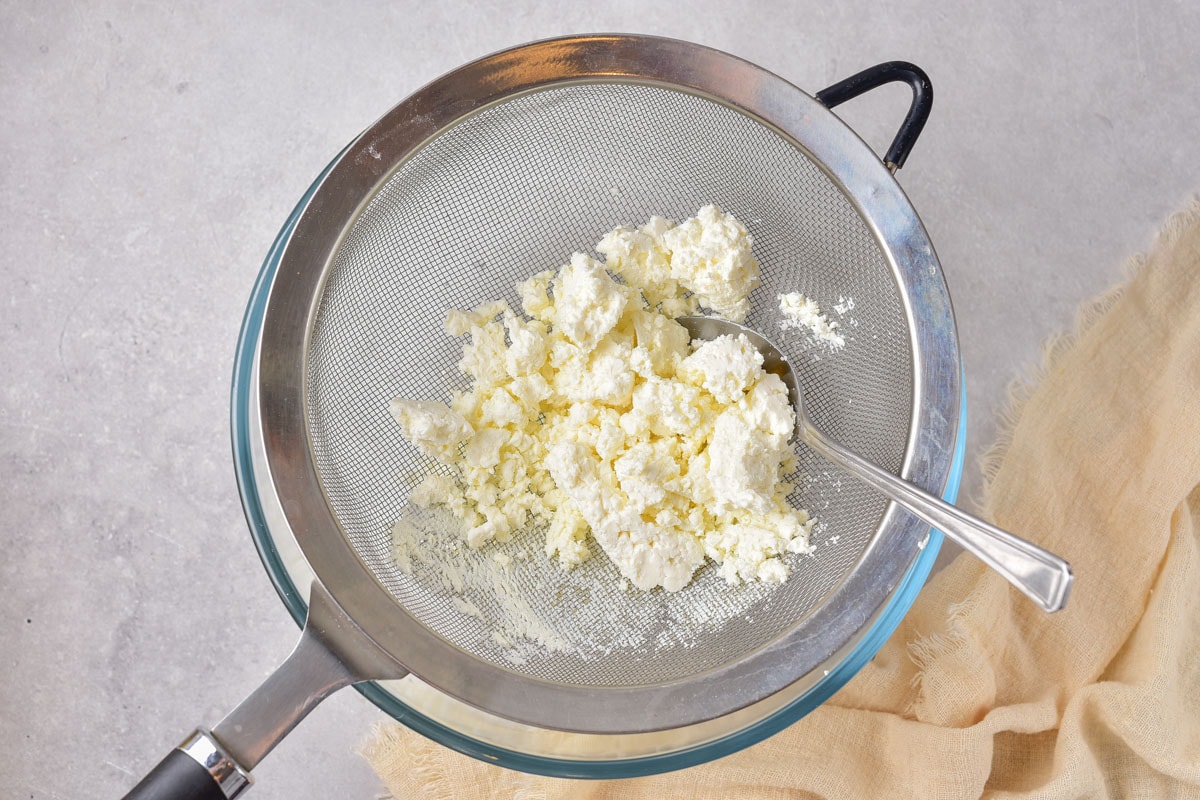 cottage cheese sitting in a metal strainer over top a bowl on a grey counter top.