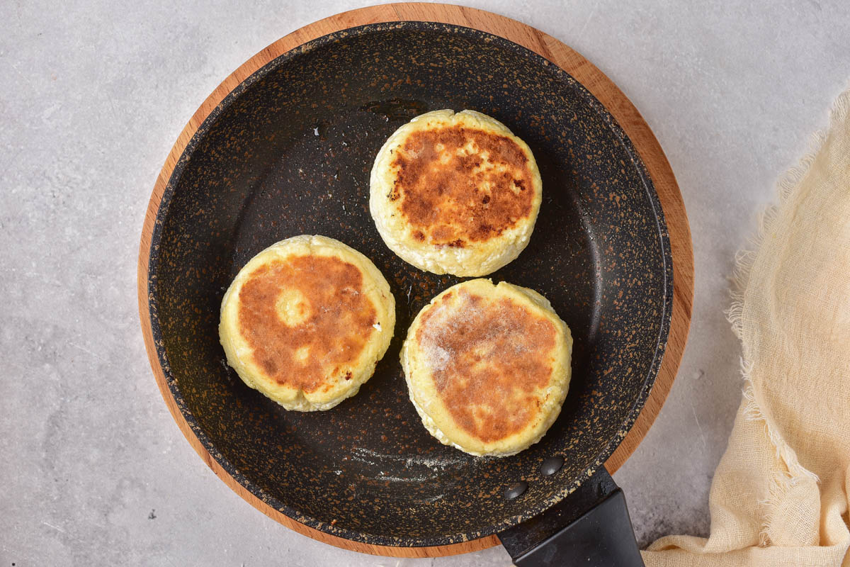 three golden brown syrniki pancakes cooking in black pan with grey counter top around.