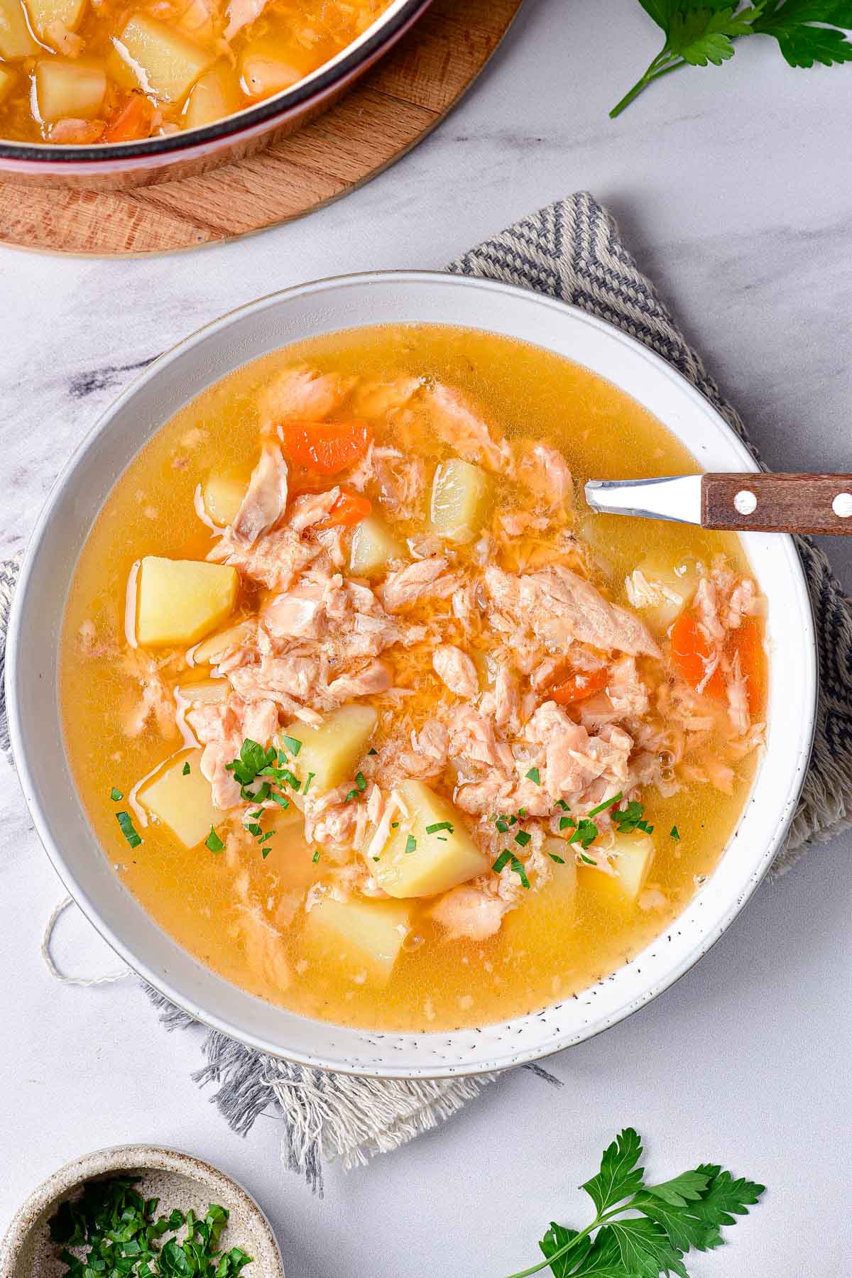 yellow fish soup with salmon and vegetables sitting in white bowl on grey cloth with spoon sticking out.