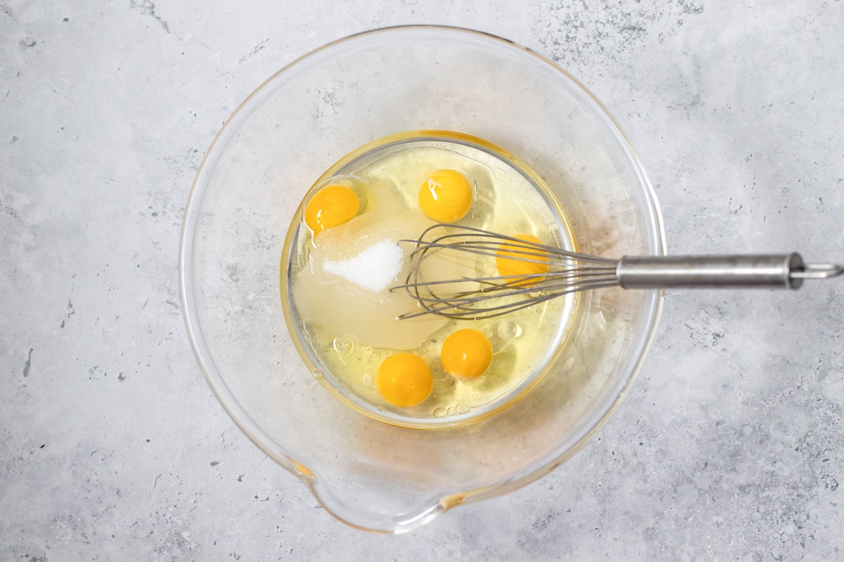raw eggs sitting in clear glass mixing bowl with silver whisk in bowl sitting on grey counter.