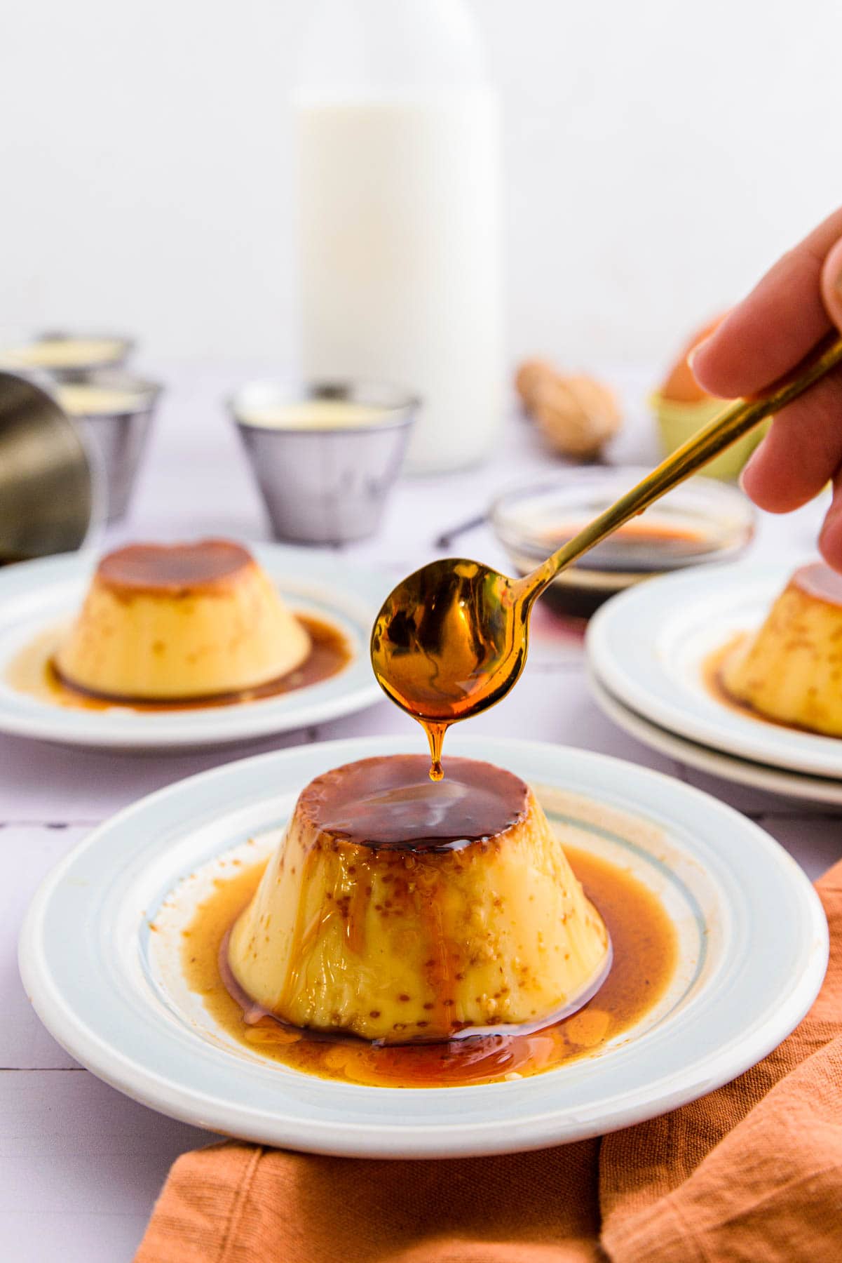 silver spoon drizzling caramel sauce on spanish flan on white plate with stuff on table behind.