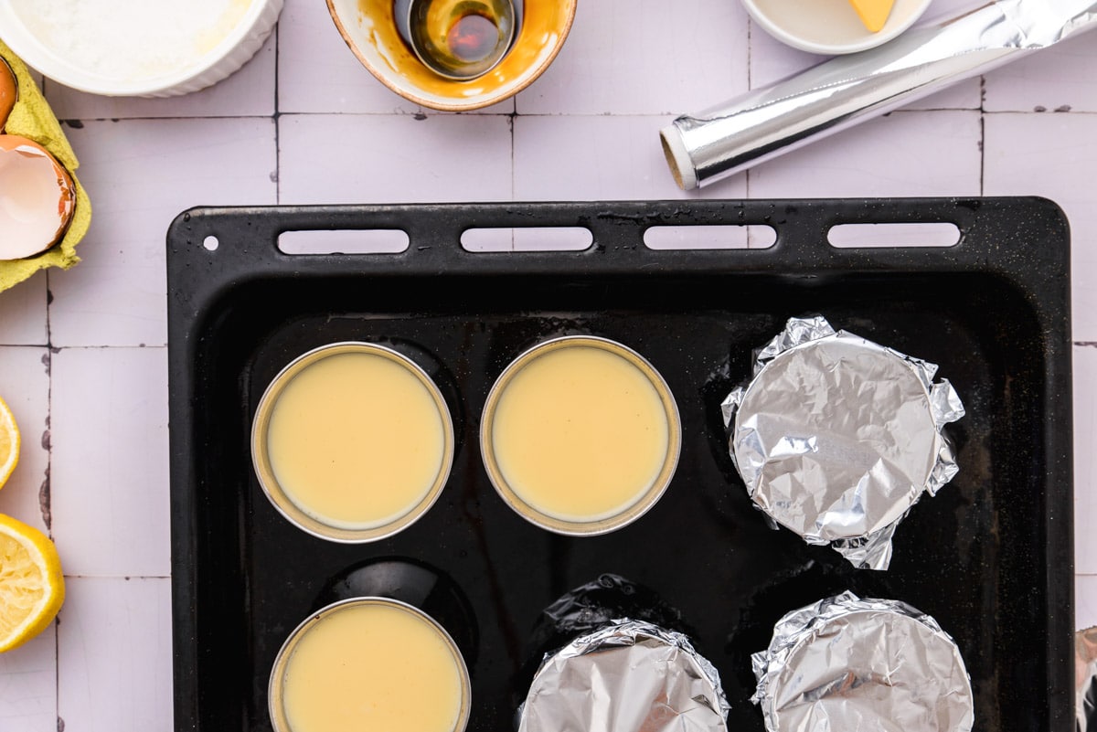 cups of flan sitting in black tray on counter some with tin foil on top.