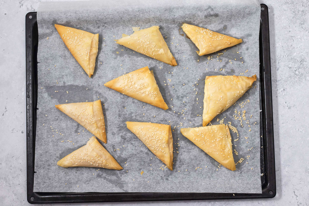 baked tiropita triangles sitting on parchment paper on black oven tray.