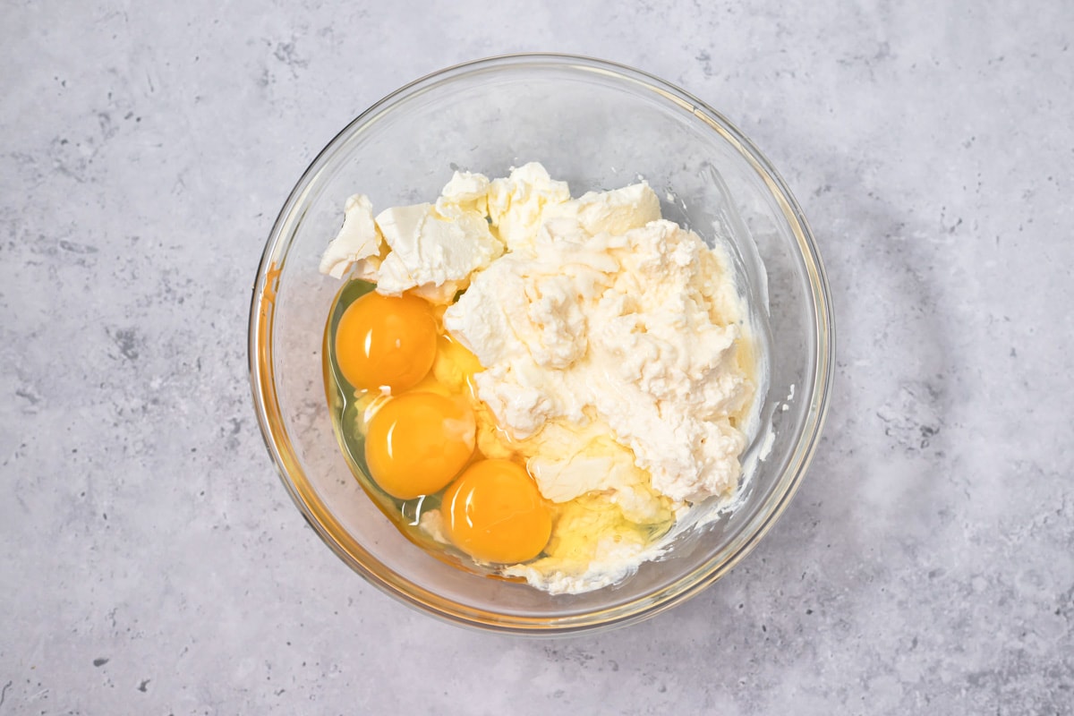 eggs and cheeses in clear glass mixing bowl on grey counter.