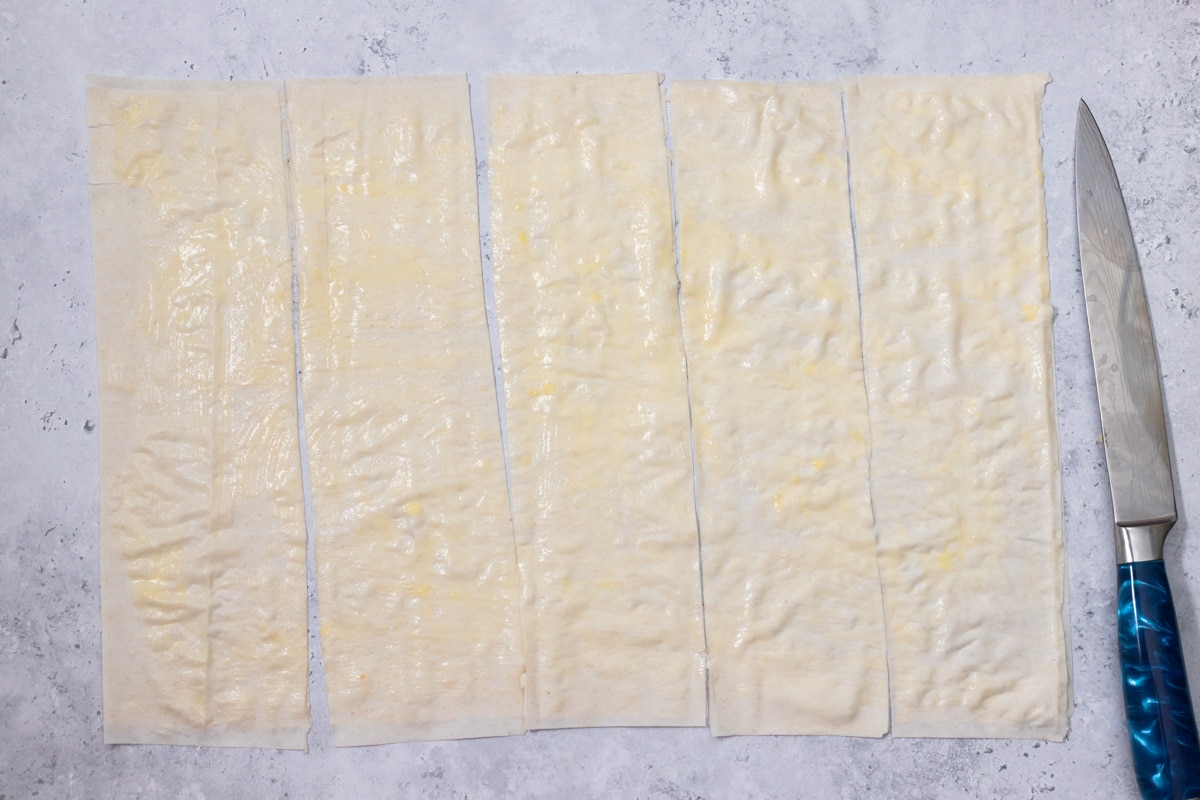 sheet of phyllo dough on counter cut into wide strips with knife beside.