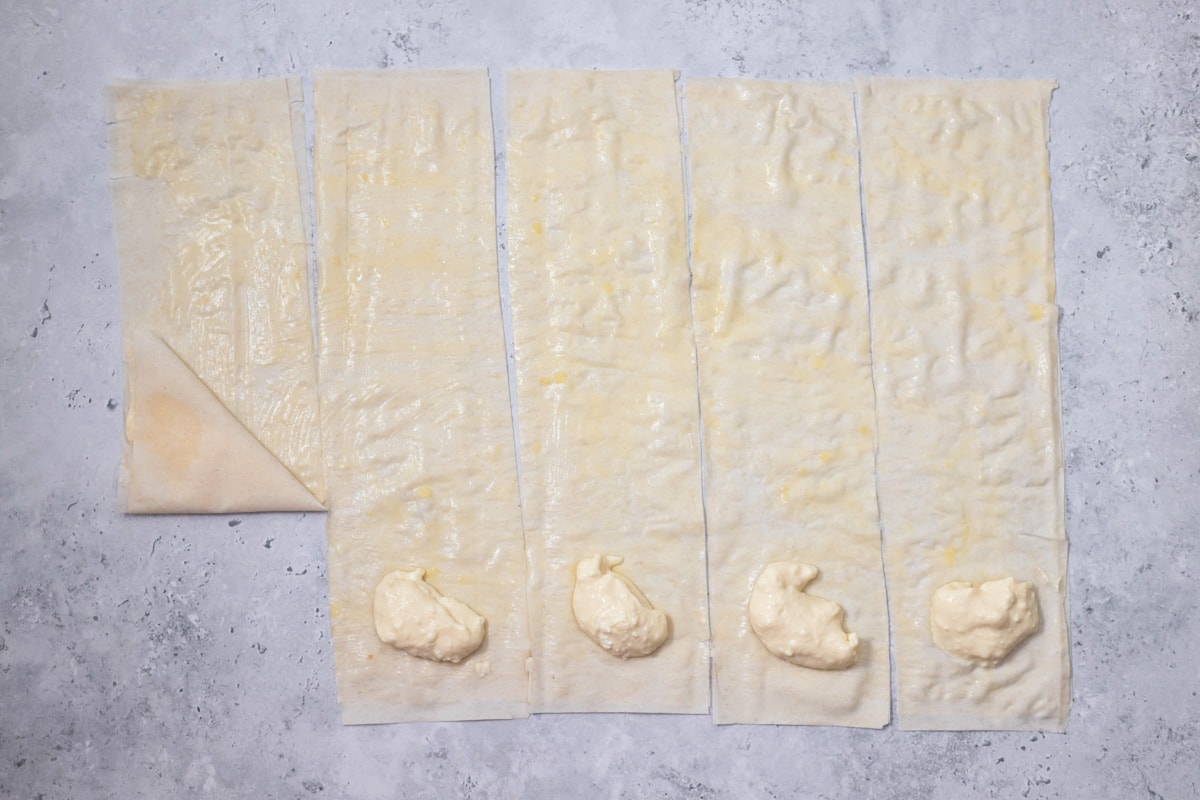 strips of phyllo dough on grey counter with spooned cheese filling at the ends with one folded into a triangle shape.