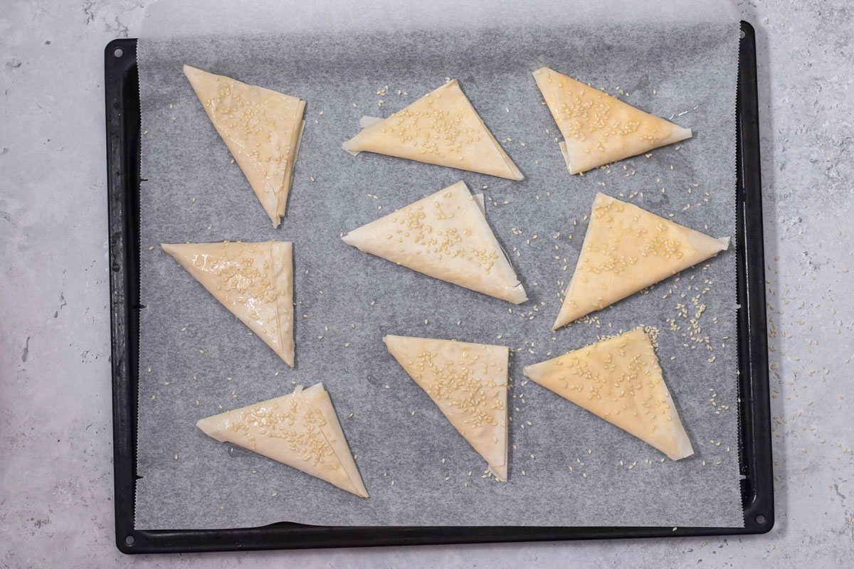 triangles of raw phyllo dough covered in sesame seeds sitting on parchment paper covered baking tray.