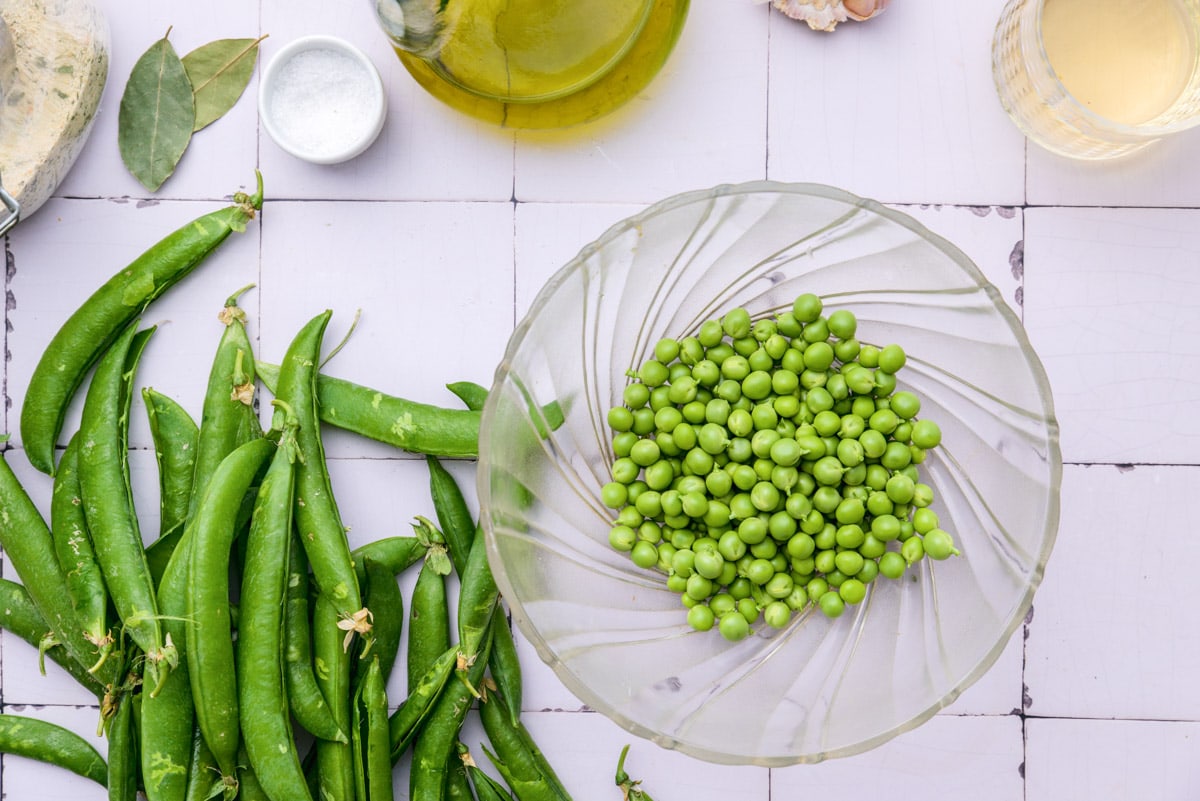 fresh green peas in clear glass bowl on counter with green pods beside.