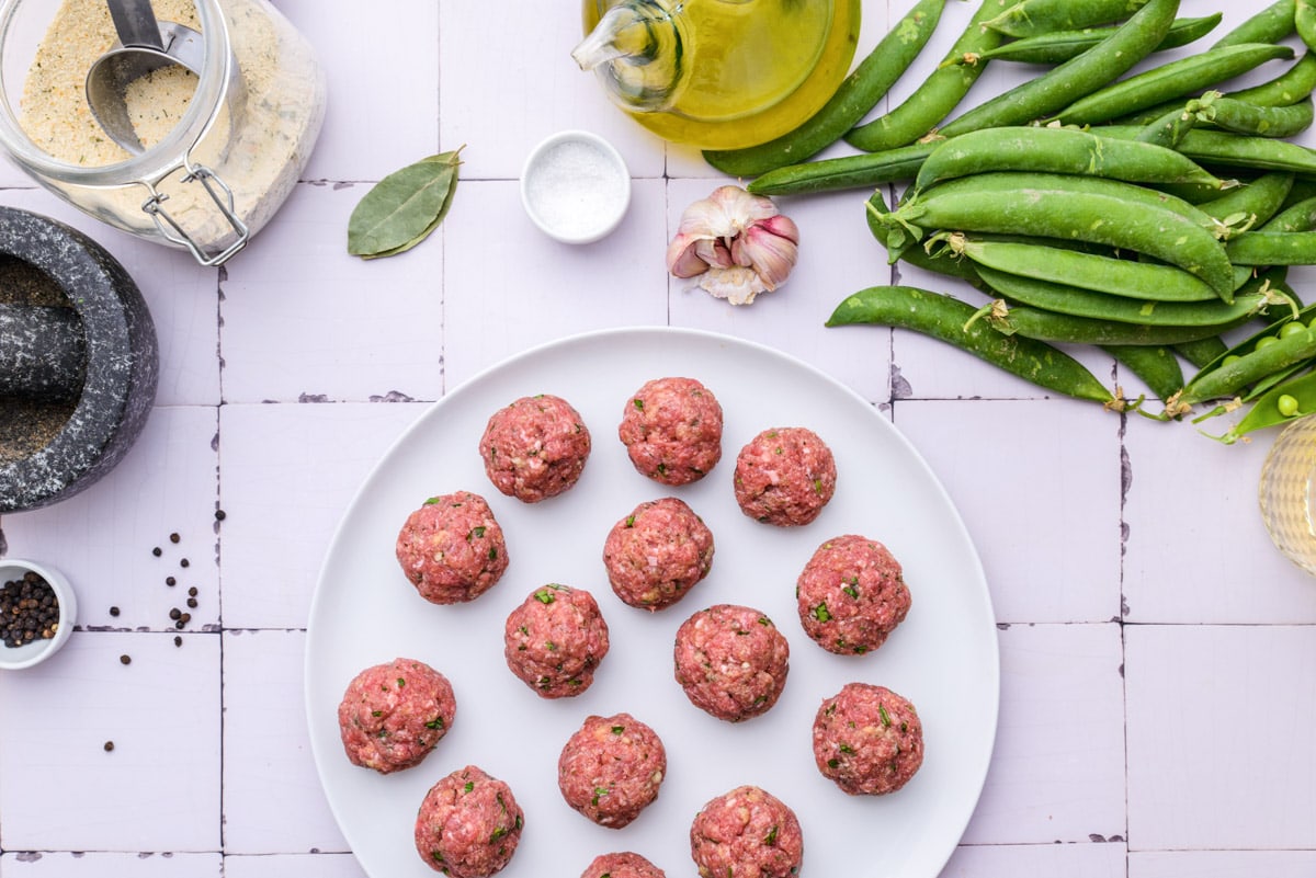 raw round meatballs on large plate sitting on counter with green peas beside.