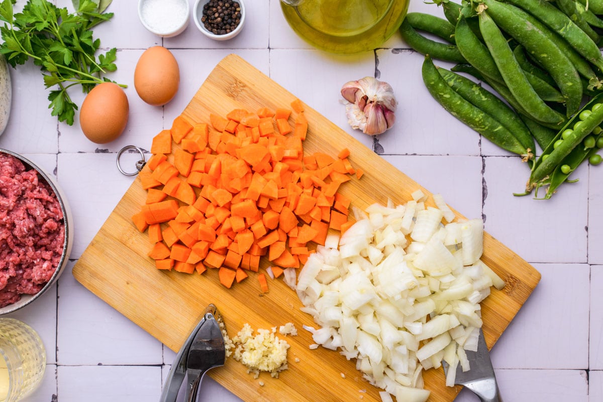 carrots and onions diced on wooden cutting board with other vegetables around on counter.