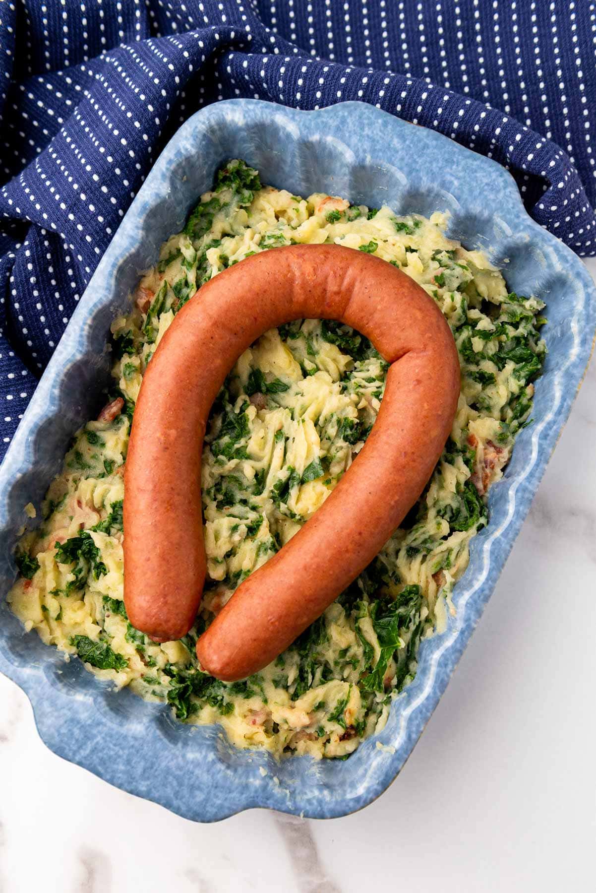 large smoked sausage sitting on top of blue baking dish filled with mashed potatoes with bacon and kale.