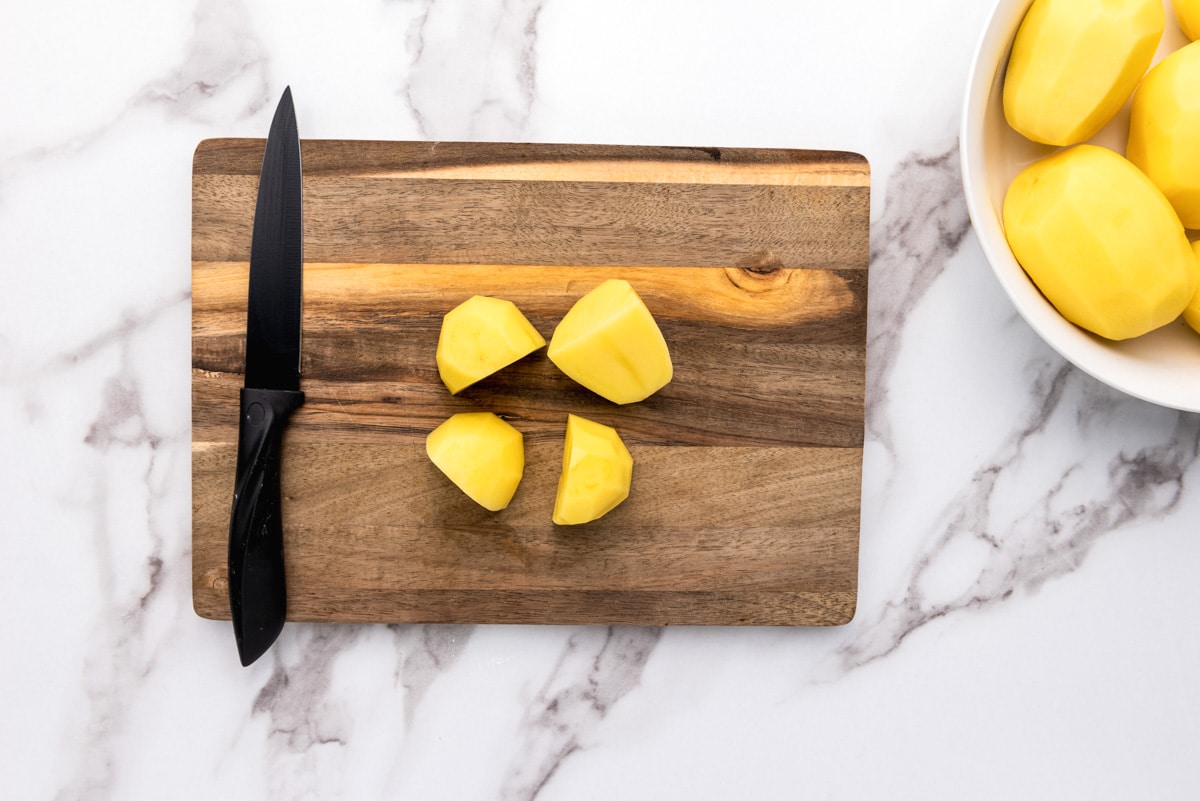 raw potato cut into quarters on wooden cutting board with black knife beside.