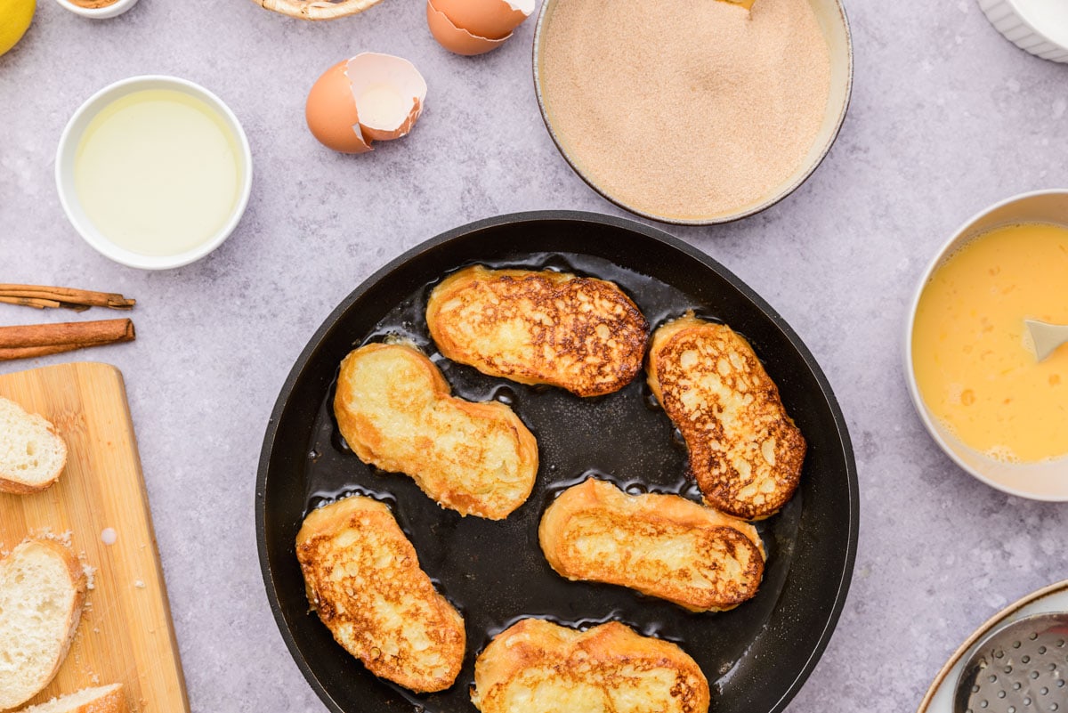 six fried spanish french toast in black pan with ingredients around.