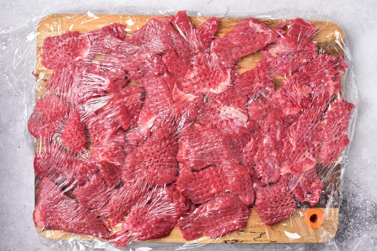 red raw beef chunks flattened under cling wrap on wooden cutting board.