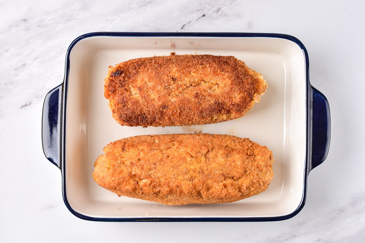 two breaded chicken kiev sitting in baking pan on marble counter.