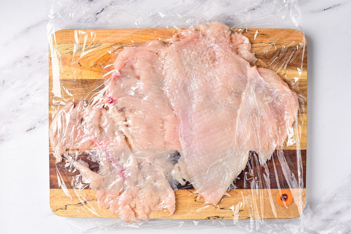 chicken breast flattened on wooden cutting board covered in cling wrap.