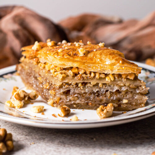 piece of baklava on plate with nuts around and brown cloth behind.