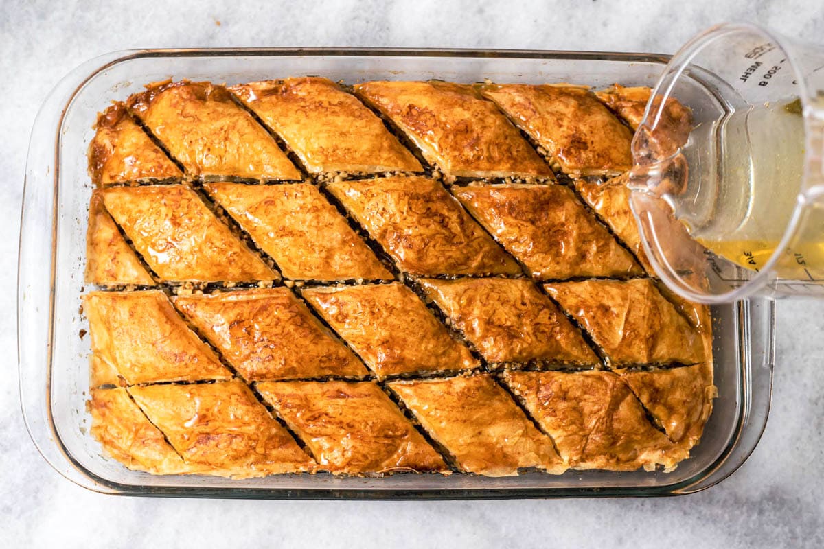 pouring hot honey syrup onto baked baklava in glass baking pan.