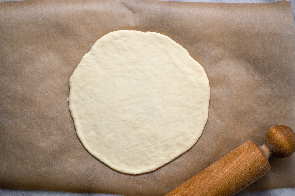 flat circle of pita cough on parchment paper with rolling pin beside.