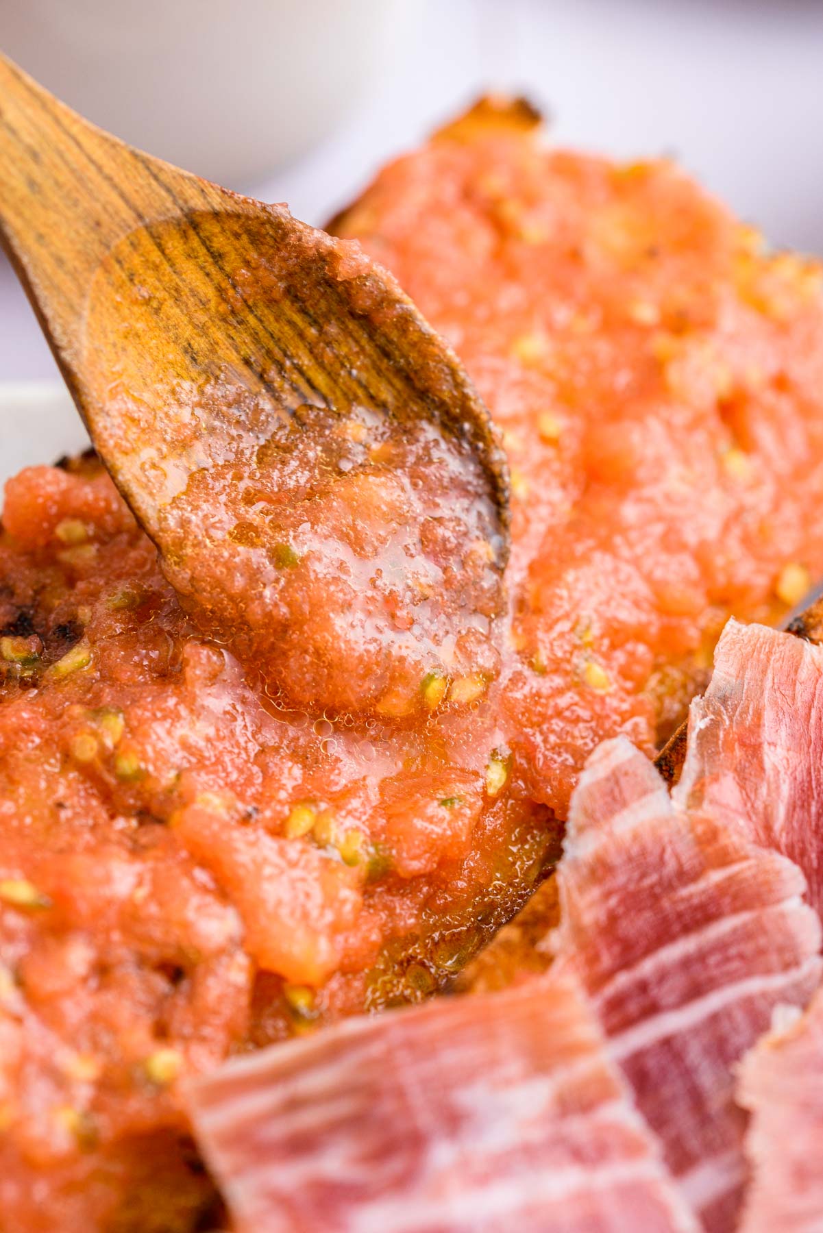 wooden spoon spreading tomato mixture with pieces of ham on bread in front.