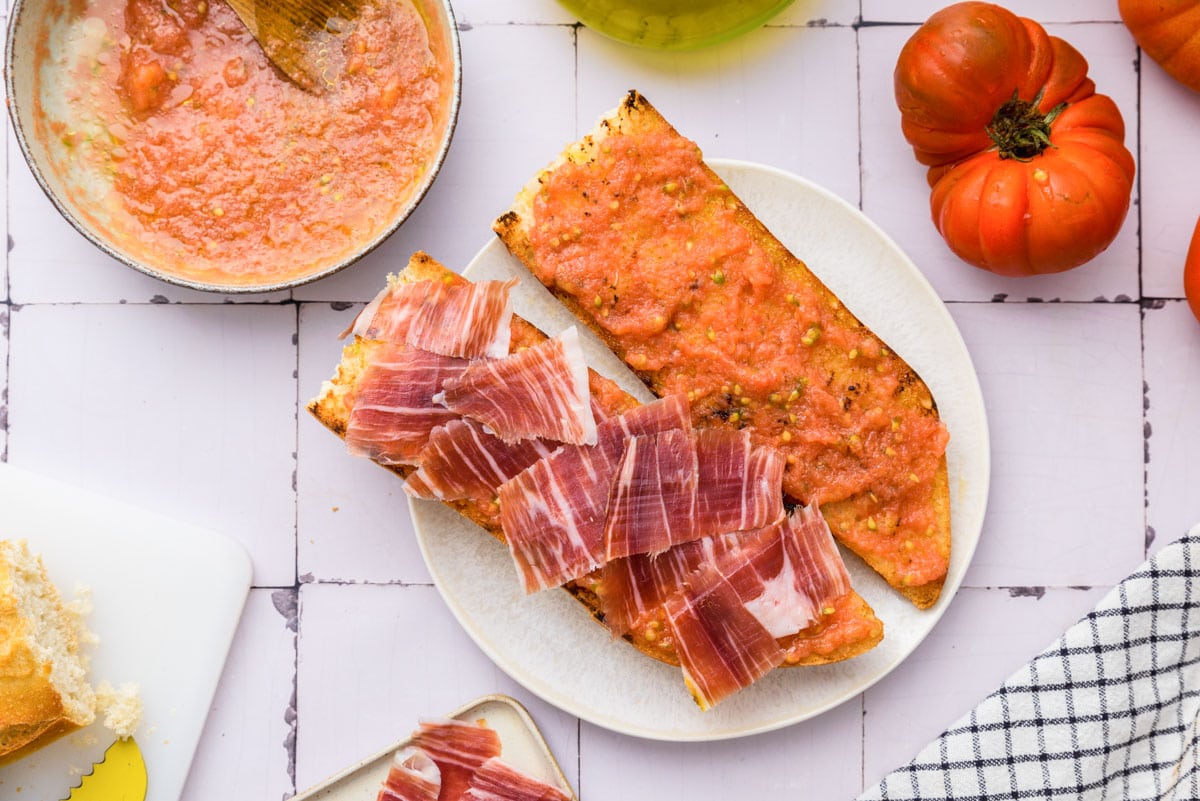 two pieces of baguette on white plate with tomato spread and one with sliced ham on top.